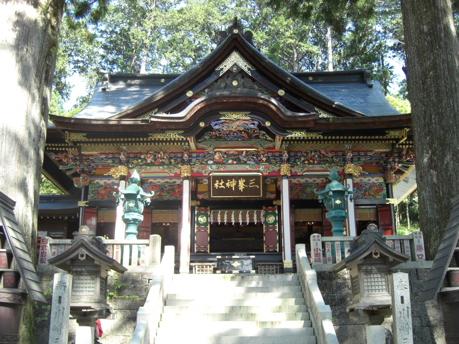 The brightly colored haiden (worship hall) of Mitsumine Shrine, surrounded by tall cedar trees